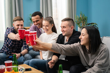 Multinational Friends having small home party, meeting, sitting on couch, clinking red cup of beer, spending, enjoying time together, watching tv