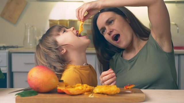 Happy Kid and mother with mango fruit in hands. Funny family crazy eating mangoes healthy dietary nutritious at home in the kitchen. Healthy lifestyle, raw food