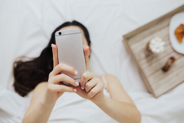 Top view of woman lying in bed with the smartphone near tray with breakfast.