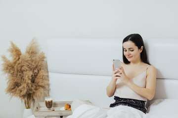 Relaxed woman eats breakfast in the bed and looks into phone.