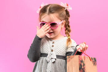 Naklejka premium Little cute girl on a pink background with a bag and money, the theme of shopping with children.
