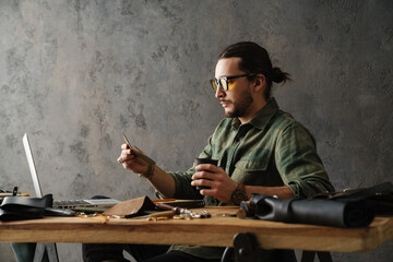 Bearded craftsman in eyeglasses drinking coffee and using laptop