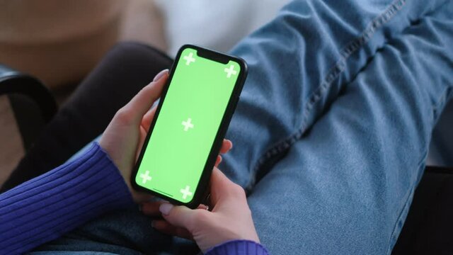 Young woman sitting on armchair at home and uses smartphone with green screen Mock-up. 4k	