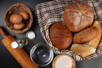 Freshly baked bread loaf bun roll round long mix verity wrapped in checkered kitchen fabric napkin towel wheat flower oil water salt eggs rolling pin slate stone over black background