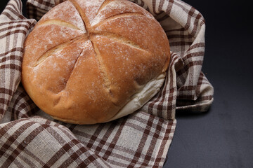 Fresh baked round bread loaf wrapped in checkered kitchen fabric napkin towel over black background