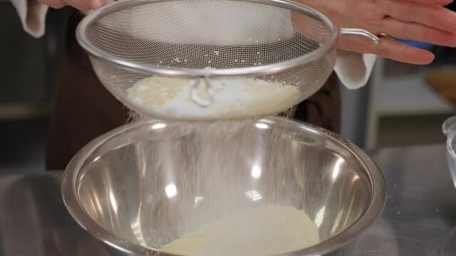 Close-up of a pastry chef sifting flour into a bowl of dough.