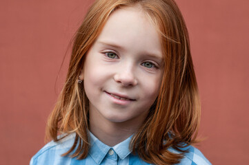Portrait of a red-haired child with green eyes 8 years old in a blue shirt on a teracot background.
