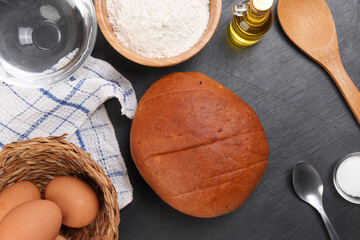 Fresh baked round bread loaf on black slate stone tablet with spoon wheat flower oil water salt kitchen napkin over white background