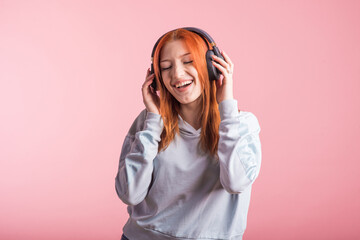 Portrait of a joyful redhead girl who listens to music on headphones in the studio on a pink...