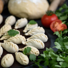 Fototapeta na wymiar Raw or uncooked dumplings on black board, cabbage and tomatoes on blurred background. Parsley or celery leaves in foreground. Close-up shot, side view. Cooking process. Soft focus.