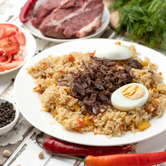 Pilaf, national oriental food. Spicy rice dish with meat. Plate of finished dish and ingredients for its preparation. Basmati rice and lamb, egg, tomato and peppers on kitchen table. Cooking process.