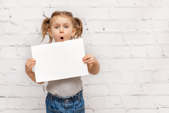 Surprised Child Over White Brick Wall Holding White Paper Sheet With Expression Face.Concept Of Advertising Goods, Seasonal Sale, Discount. Shopping And Retail. Promoting Goods. Place For Text. Mockup