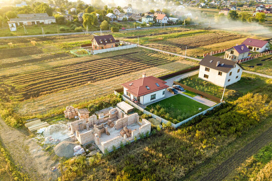 Top down aerial view of two private houses, one under construction with concrete foundament and brick walls and another finished with red tiled roof.
