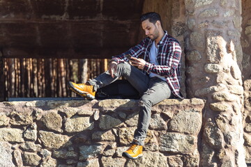 Fototapeta premium Attractive man sitting on stone wall looking at mobile phone.