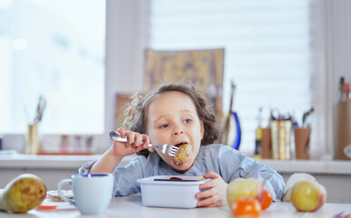 Cute small girl sitting at the table at home kindergarten or school class and eating a lunch with big pleasure from blue lunchbox using fork. Happy child eating tasty food concept. High quality photo