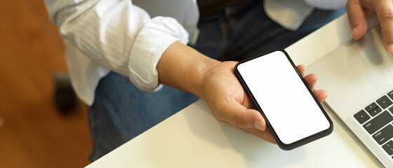 Top view of male freelancer holding smartphone with mock up screen in his hand on worktable