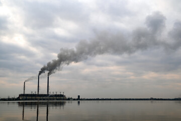 Coal power plant high pipes with black smoke moving upwards polluting atmosphere with reflections of it in lake water.