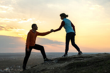 Man and woman hikers helping each other to climb stone at sunset in mountains. Couple climbing on high rock in evening nature. Tourism, traveling and healthy lifestyle concept.