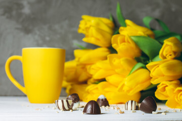 Chocolate candies, cup and bouquet of yellow tulips on white wooden surface on gray background. Copy space