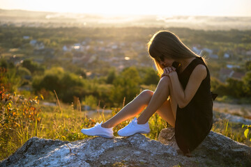 Pretty sad woman in black short summer dress sitting on a rock thinking outdoors at sunset....