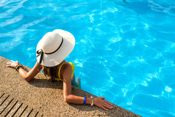 Top down view of young woman wearing yellow straw hat resting in swimming pool with clear blue water on summer sunny day.