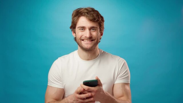 Young man looks at cell phone screen with joy, recieved good news, against blue background