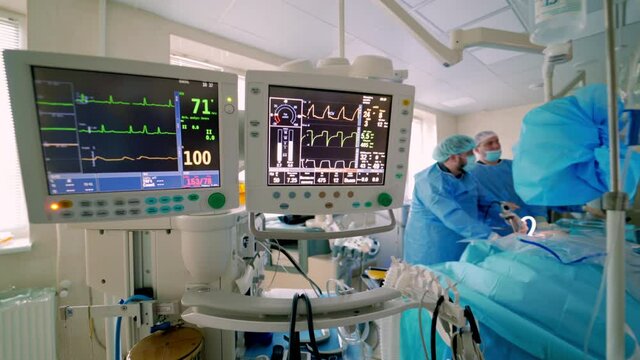 Hospital monitor in the operating room. Screen shows vital signs of a patient on the background of surgical procedure. Teamwork of doctors.
