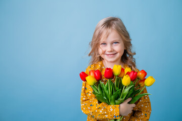 Cute little blonde girl holding a bouquet of tulips in a cotton dress on a blue background. spring,...