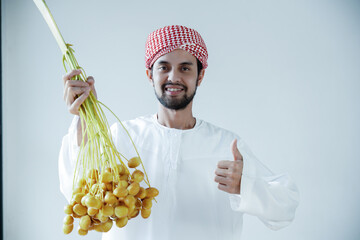 Young Arab man with beard is holding bunch of fresh date palms. Muslim farmer smiling and looking at camera with pride and thumb up. White background