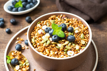Homemade organic buckwheat granola with blueberries, hazelnuts, peanuts, nutmeg, pumpkin seeds and flax in a ceramic bowl on a brown background. Healthy vegetarian breakfast closeup