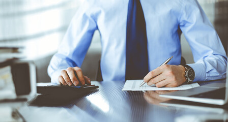 Unknown man accountant in blue shirt use calculator and computer with holding pen on while staying at home during covid pandemic. Taxes and audit concept