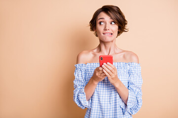 Photo portrait of anxious woman biting lower lip looking at blank space holding phone in two hands isolated on pastel beige colored background