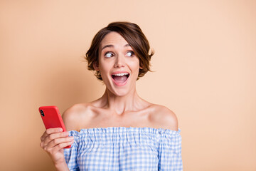 Photo portrait of excited girl looking at blank space holding phone in one hand isolated on pastel beige colored background