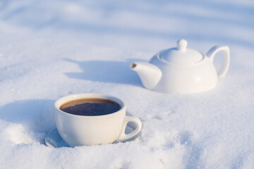 White cup of hot tea and teapot on a bed of snow and white background, close up