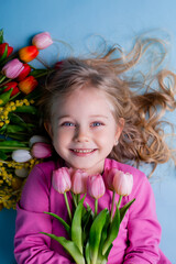 Obraz na płótnie Canvas A cute little girl lies among the tulips on a blue background in the studio. spring concept, text space