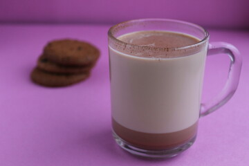 a cup of hot chocolate cocoa in a glass cup in the background lies homemade cookies and cinnamon sticks on a lilac purple background with a place for text and copyspace