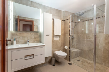 Beige colored bathroom interior, a shower with a glass door and a ceramic toilet, also a few towels on the walls.