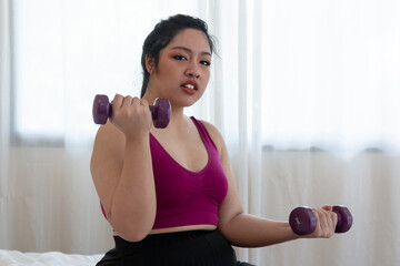 Fototapeta na wymiar Young Overweight woman doing exercise with dumbbells at home. Overweight fat woman with measure tape on neck holding dumbbell in hands while sitting at home. Woman exercise weight loss at home.