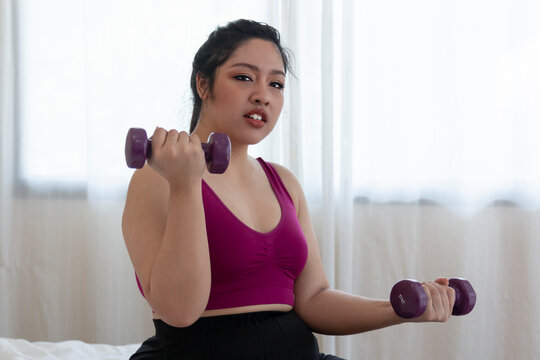 Young Overweight woman doing exercise with dumbbells at home. Overweight fat woman with measure tape on neck holding dumbbell in hands while sitting at home. Woman exercise weight loss at home.