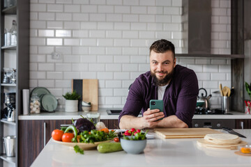 Fototapeta na wymiar Happy Man smiling at Camera and holding a Smartphone and searching online recipes on food app with fresh healthy vegetables and cutting board on the table, copy space