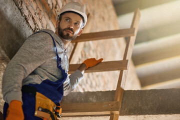 Portrait of handsome male builder in blue overalls and hard hat looking away, climbing down the...