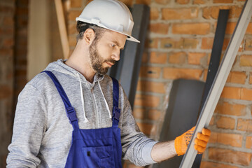 Portrait of focused male builder wearing overalls and hard hat holding a metal stud for drywall on interior site building