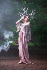 Full Length Portrait of Tranquil Caucasian Girl Posing In Light Dress With Artistic Deer Horns In Summer Forest With Grey Smoke Behind.
