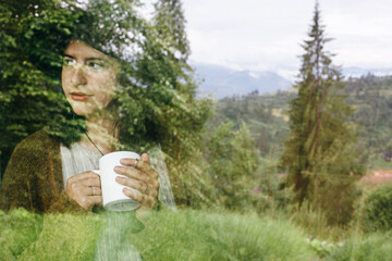 Reflection of young woman with morning coffee or tea in window with view on mountains and trees