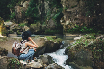 Travel and wanderlust. Stylish woman traveler with backpack sitting at river in mountains. Tranquil