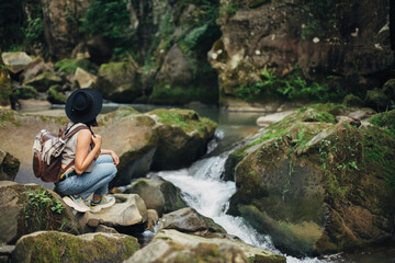 Stylish woman traveler with backpack sitting at river in mountains, relaxing. Travel and wanderlust