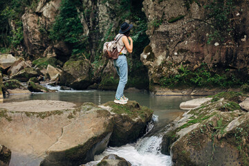 Stylish woman traveler with backpack relaxing at river in mountains. Travel and wanderlust