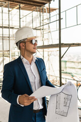 Man architect wearing formal suit and hard hat during building construction control holding a blueprints on a construction site