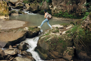 Stylish woman traveler with backpack walking on rocks of river in mountains.Travel and wanderlust