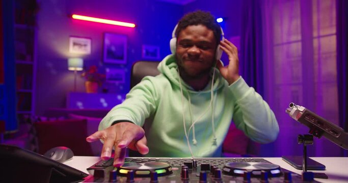 Camera approaching emotional happy African American young male dj playing music on mixer console sitting in neon lights at home recording studio. Close up of dj scratching vinyl creating beats